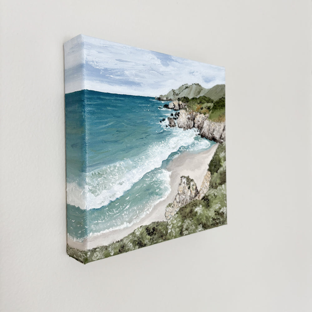 Original oil on canvas painting of a coastal landscape with blue ocean and green cliffs, hanging on a white wall. 