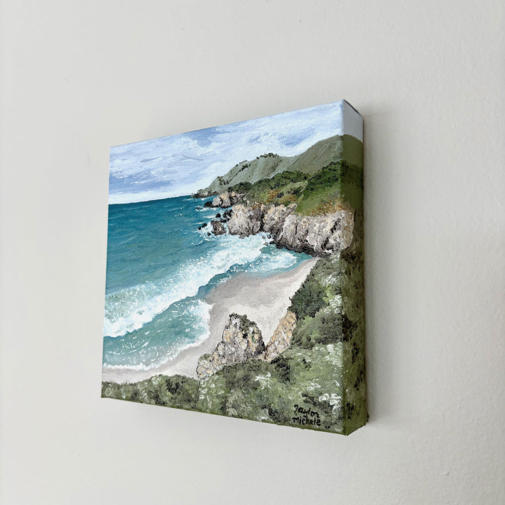 Original oil on canvas painting of a coastal landscape with blue ocean and green cliffs, hanging on a white wall. 