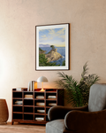 Framed artwork of a coastal scene hanging on a wall above a bookshelf and armchair.
