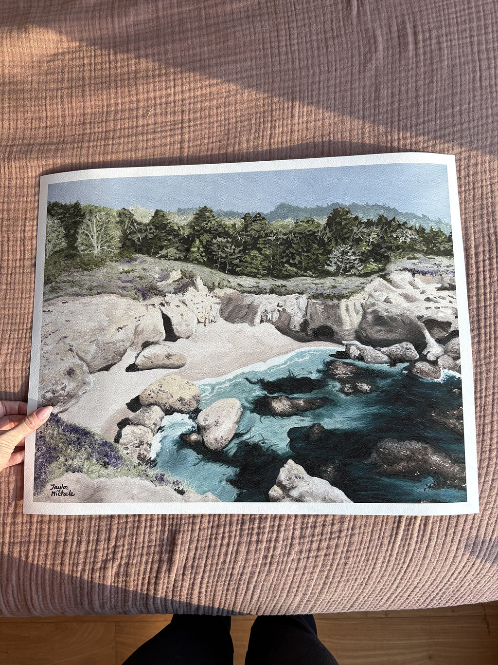 Woman's hand holding a fine art print of a Carmel Highlands rocky coastal beach cover nature scene with rocks and water on a earthy pink fabric background