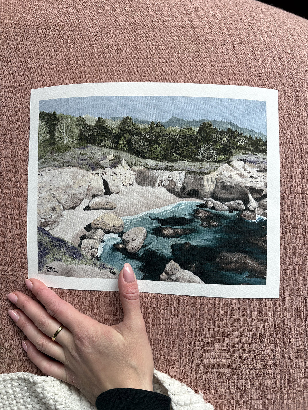 Hand holding a photograph of a natural landscape with rocks and water on a textured pink surface.