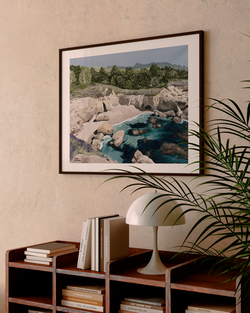 Framed landscape print on a wall above a bookshelf with a lamp and books.