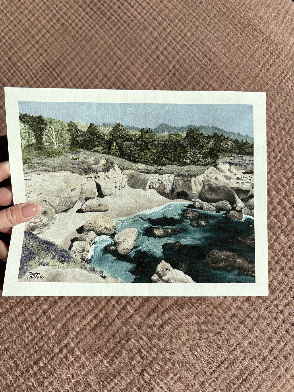 a woman's hand holds up a 8x10" mini fine art print with a scenic view of a rocky coastal cove ocean landscape held against a textured sandy beach