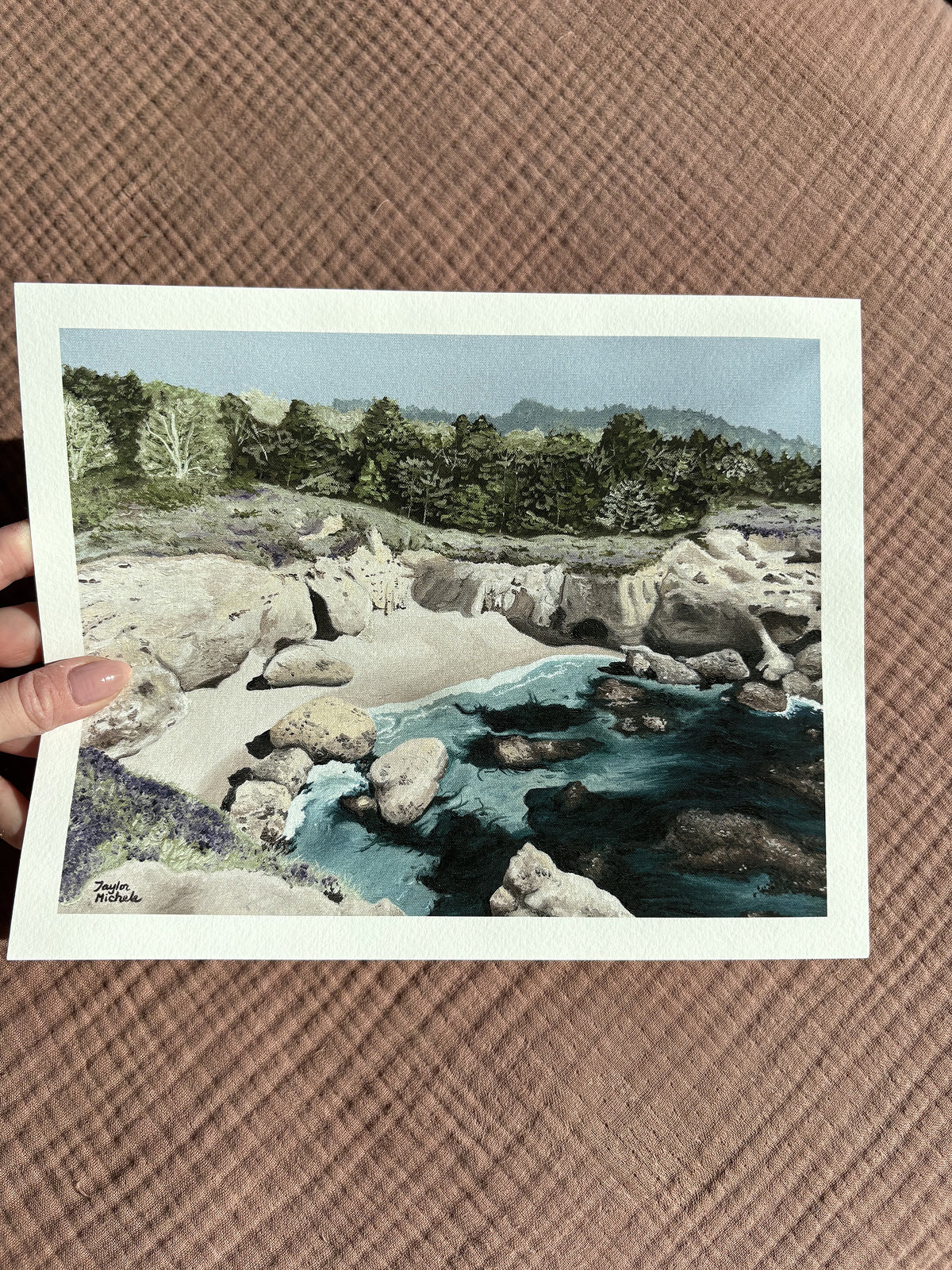 a woman's hand holds up a 8x10" mini fine art print with a scenic view of a rocky coastal cove ocean landscape held against a textured sandy beach