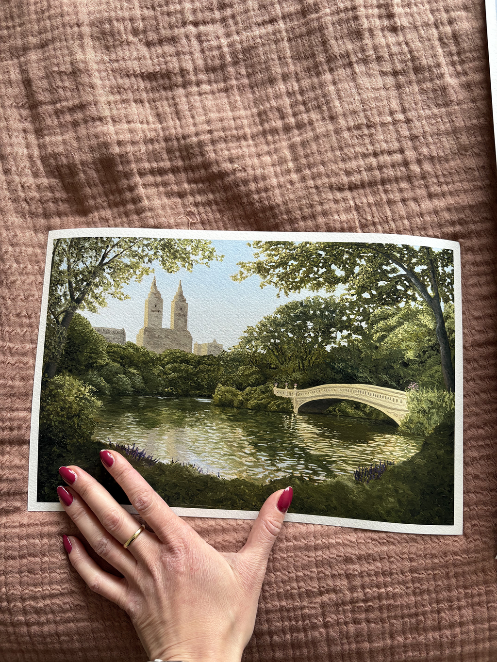 Woman's hand holding a 18 x 12 in unframed Central Park print. This shows with a scenic illustration of Central park with the bow bridge and trees.