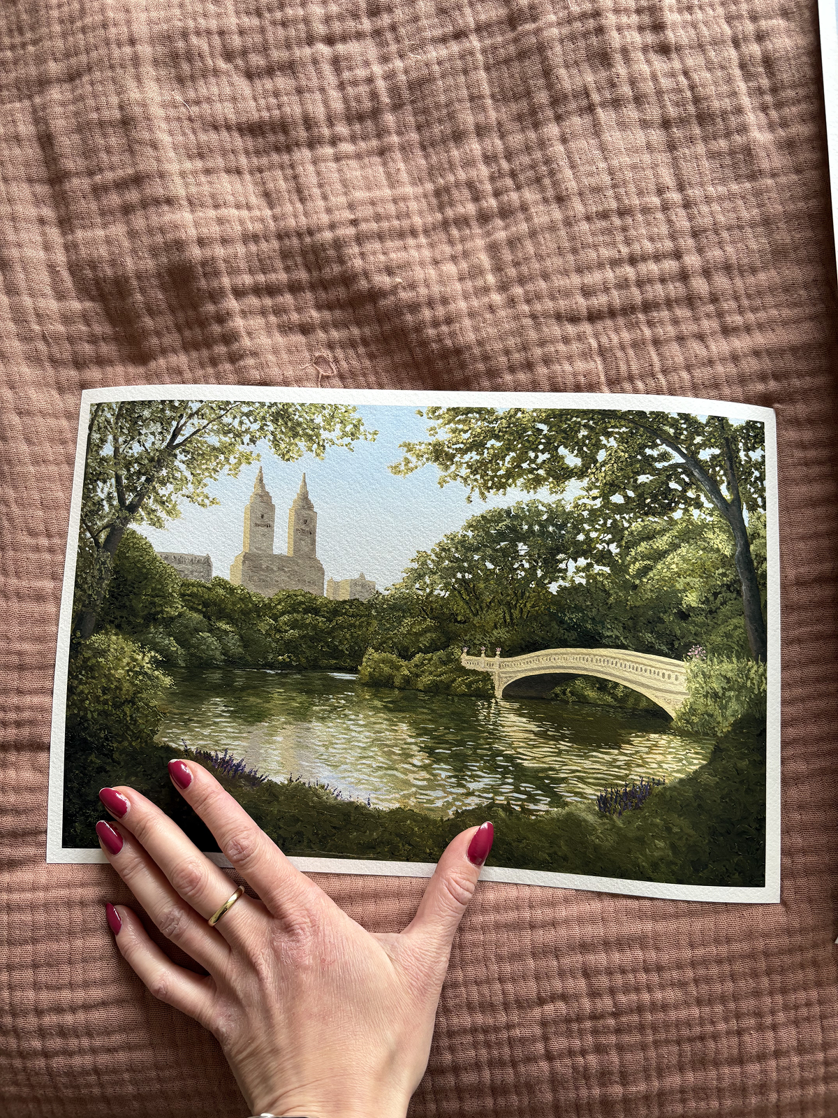 Woman's hand holding a 18 x 12 in unframed Central Park print. This shows with a scenic illustration of Central park with the bow bridge and trees.