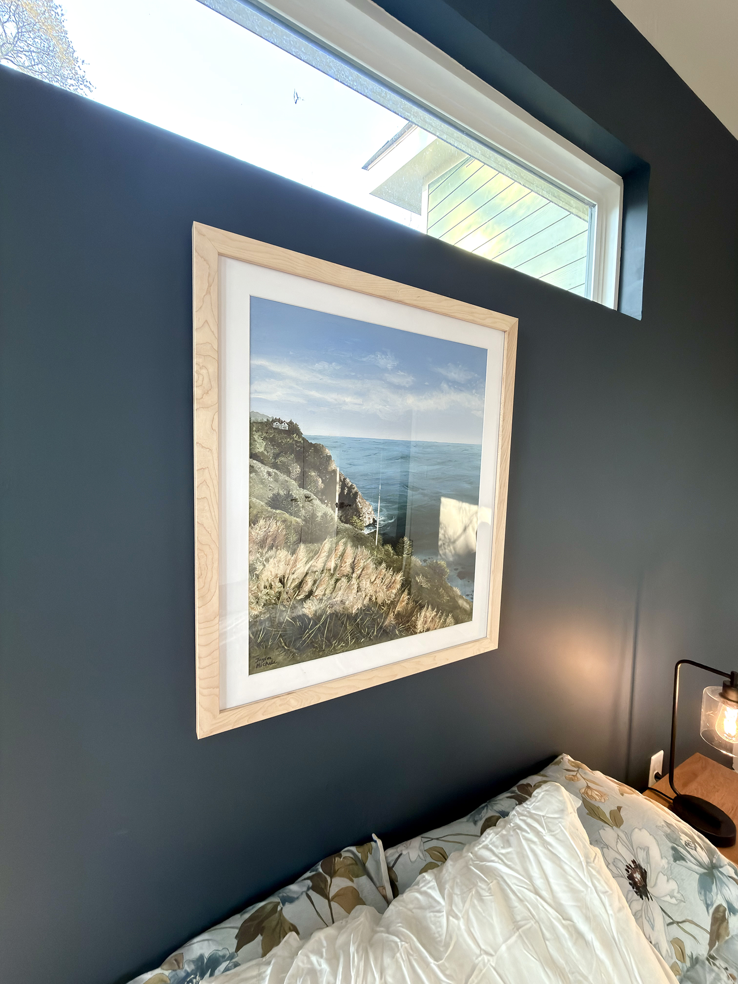 Framed artwork of a coastal scene on a dark blue wall above a bed.