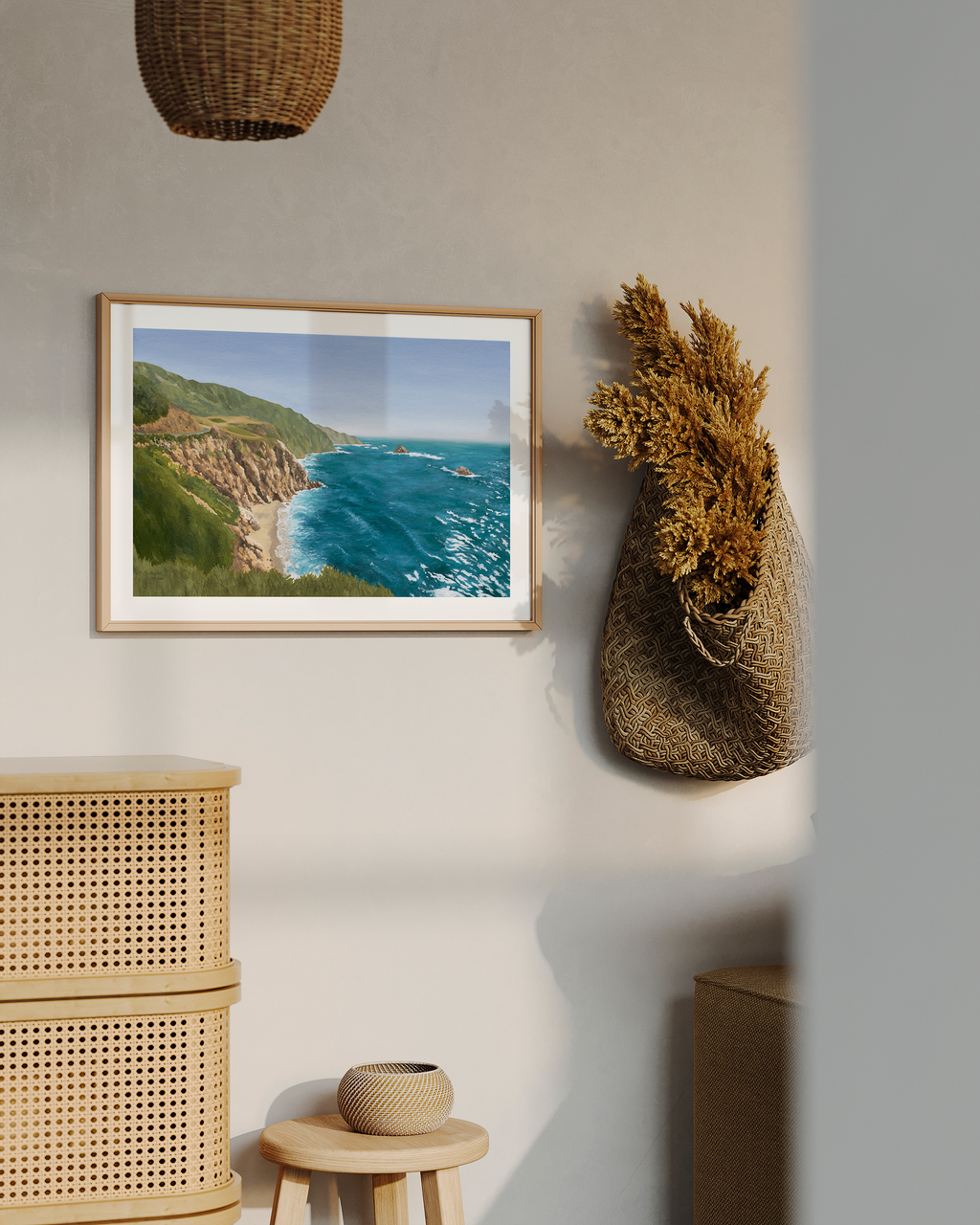 Framed California coastal landscape artwork displayed in a warm minimalist interior with woven textures and natural decor. Original oil painting captures the dramatic Big Sur coastline with rugged cliffs, turquoise ocean waves, and soft coastal light.