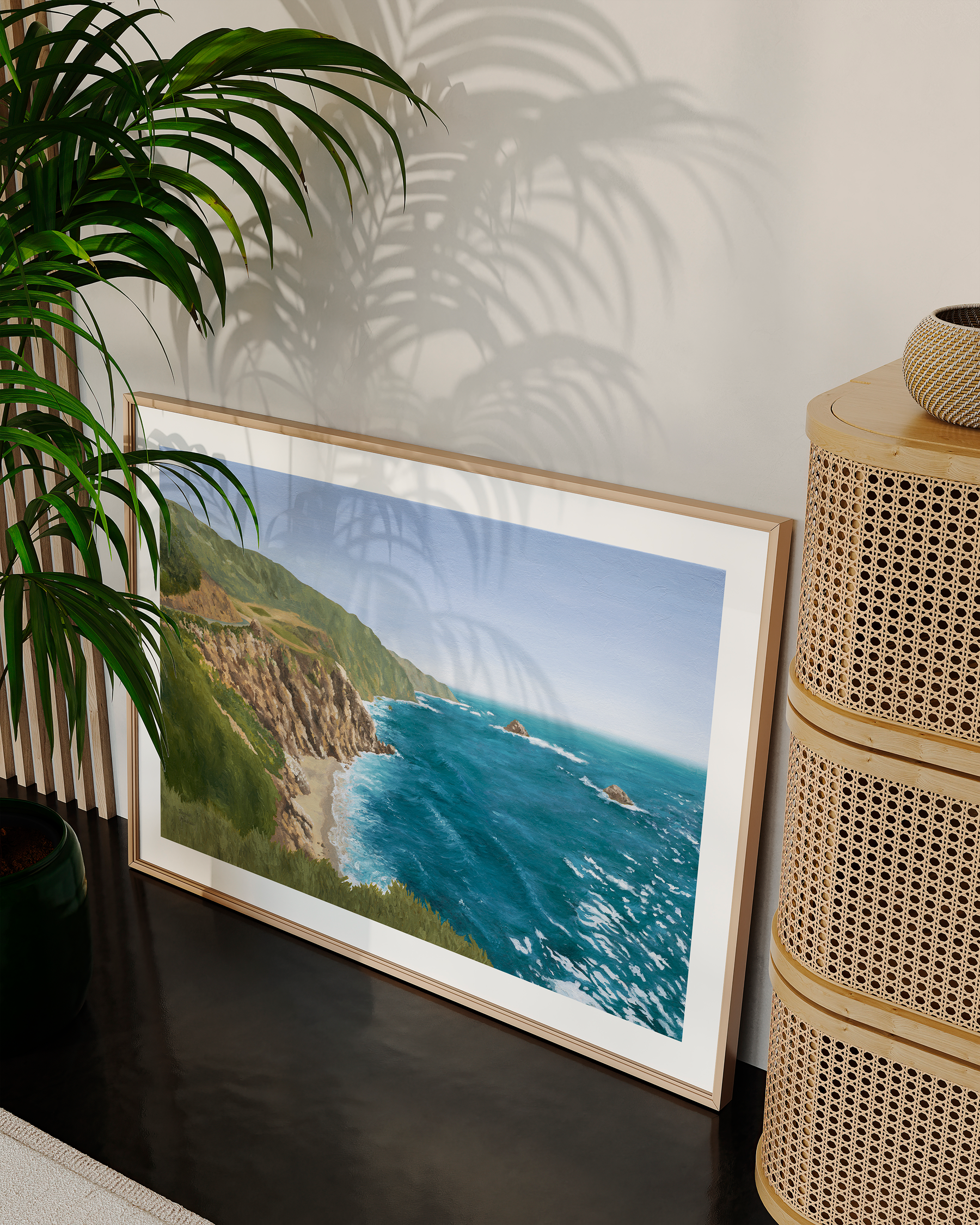 Framed coastal California landscape oil painting featuring dramatic ocean cliffs, turquoise blue water, and rugged shoreline inspired by the Pacific Coast Highway. Fine art print styled in a warm neutral living room with a leafy green plant, natural textures and woven dresser.