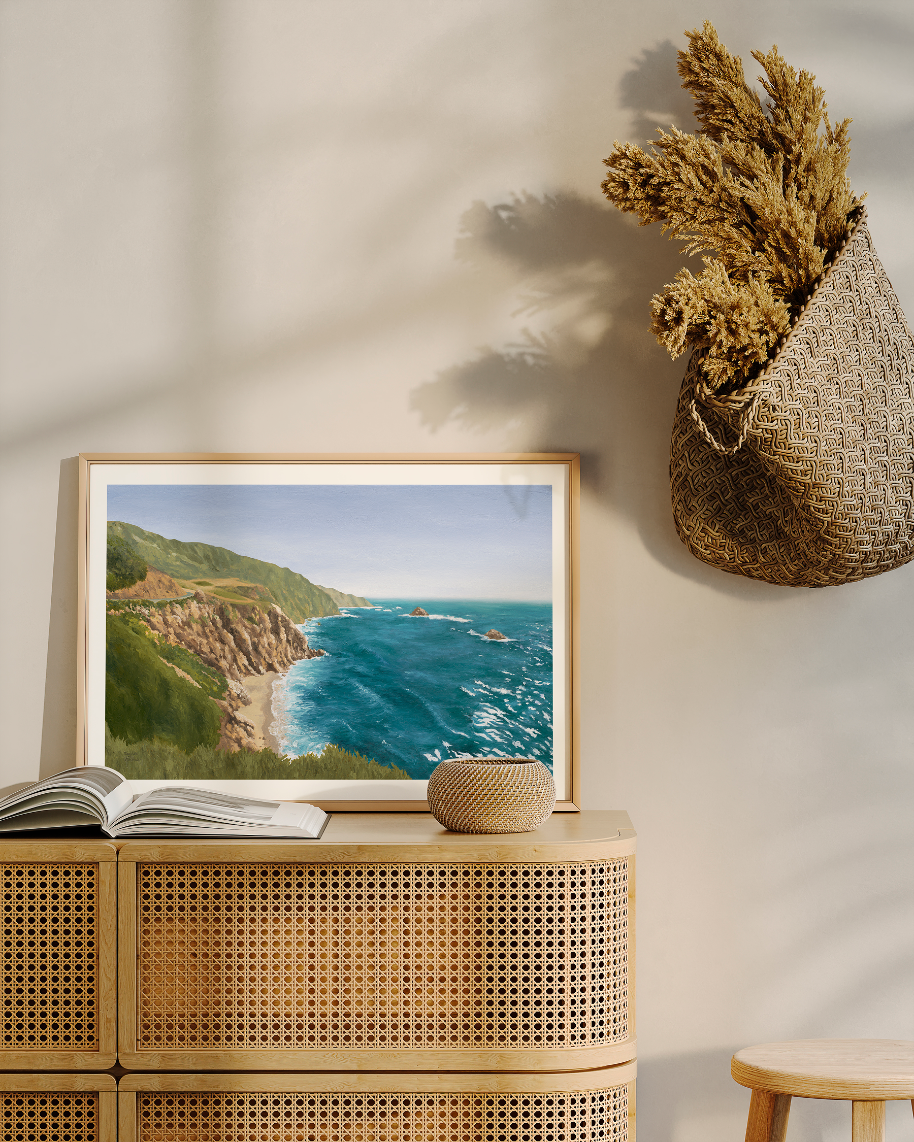 Framed California coastal landscape painting displayed on a woven cane cabinet in a warm neutral interior. Original oil painting features rugged ocean cliffs, turquoise Pacific water, and soft coastal light inspired by the Big Sur coastline.