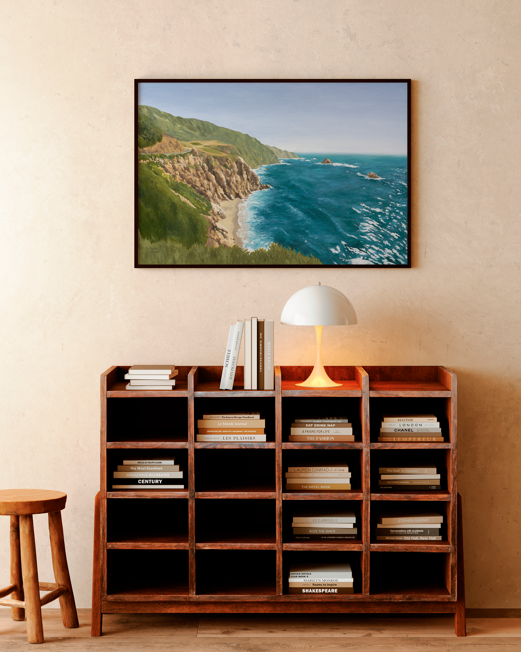 Large framed coastal California oil painting hung above a mid-century wooden bookshelf in a warm minimalist interior. Artwork features rugged Big Sur cliffs, turquoise ocean waves, and sunlit Pacific coastline with natural earthy tones.