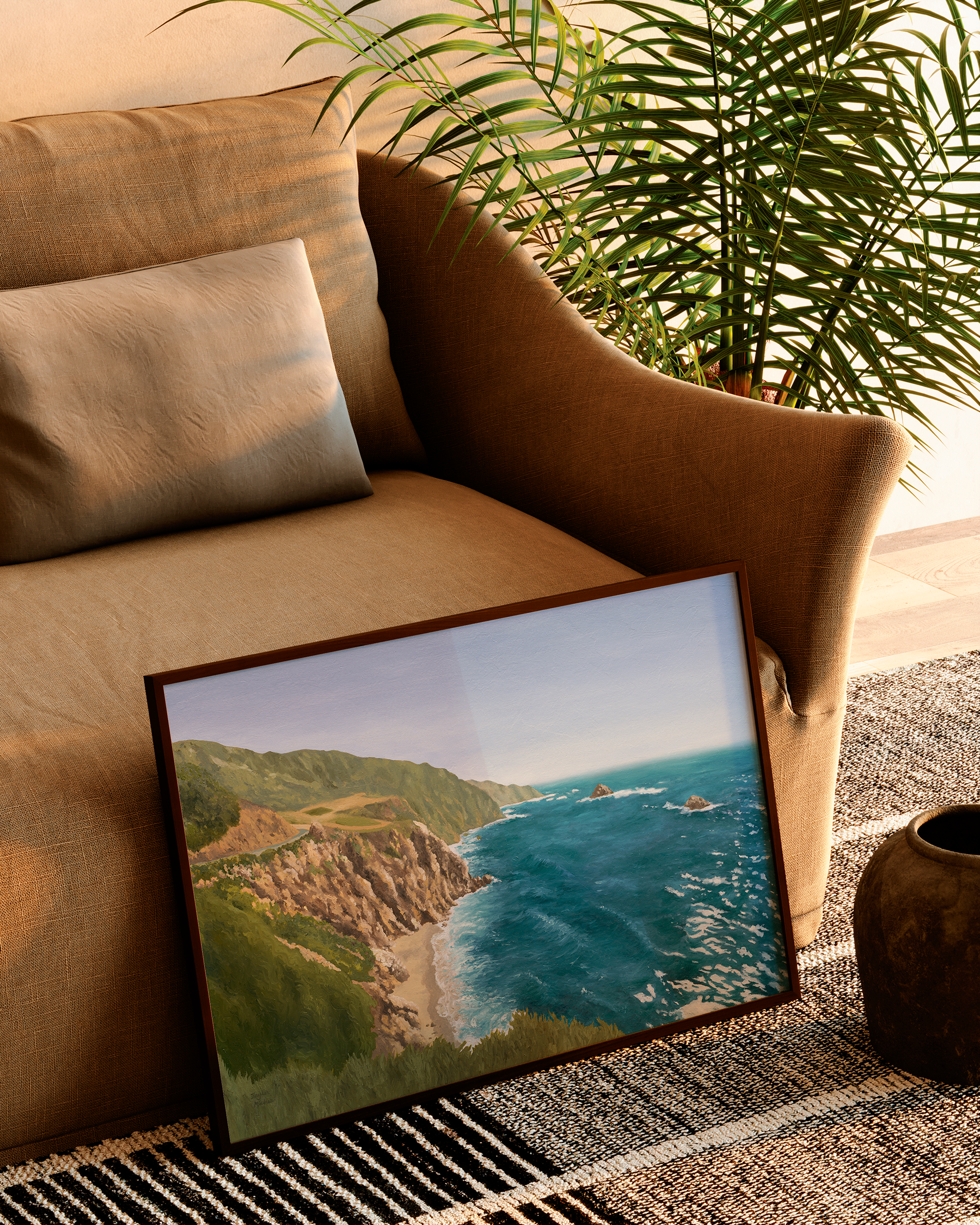 Framed Big Sur coastal landscape painting leaning against a warm neutral sofa beside a palm plant in a sunlit living room. Original California oil painting features rugged cliffs, turquoise Pacific water, and soft natural coastal scenery.