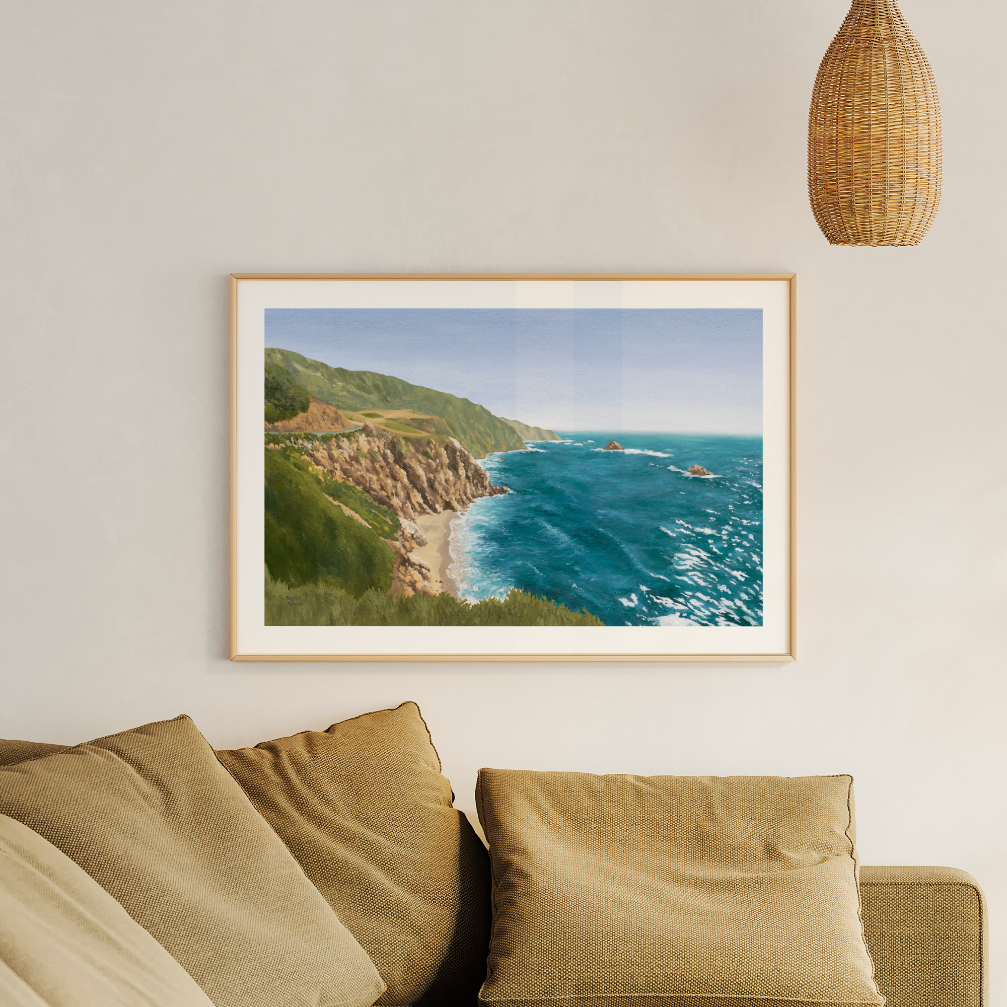 Framed coastal California landscape oil painting featuring dramatic ocean cliffs, turquoise blue water, and rugged shoreline inspired by the Pacific Coast Highway. Fine art print styled in a warm neutral living room with natural textures and woven pendant lighting.