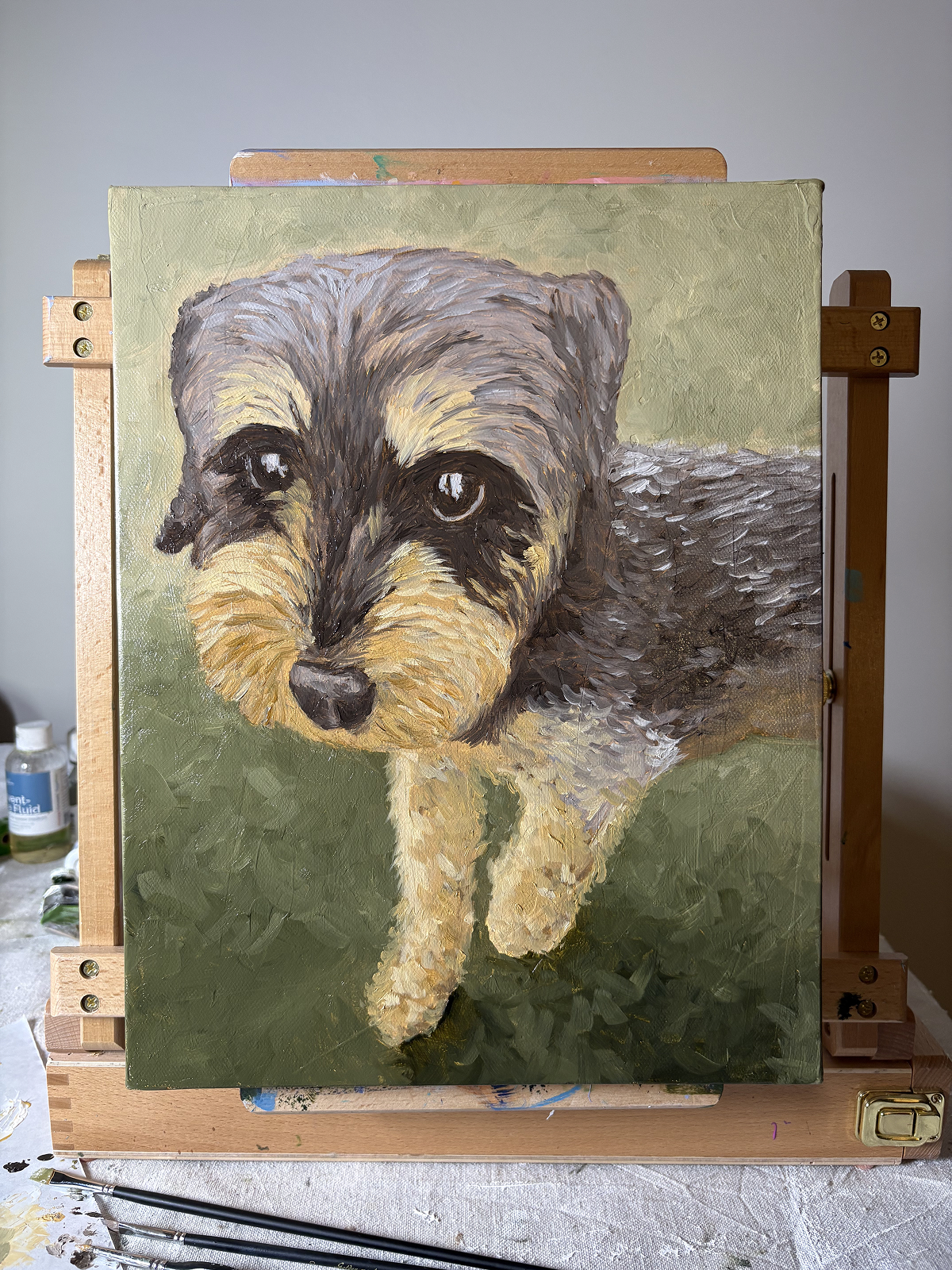 Small oil painting portratit of a black, gray and tan mini schnauzer dog with a soft green background. The painting sits on an easel with art supplies in the background.