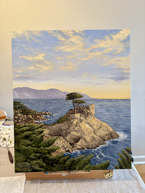 Canvas painting of a coastal landscape with a lone cypress tree on a rocky coast in Pebble Beach, CA, displayed on a table top easel on top of a table.