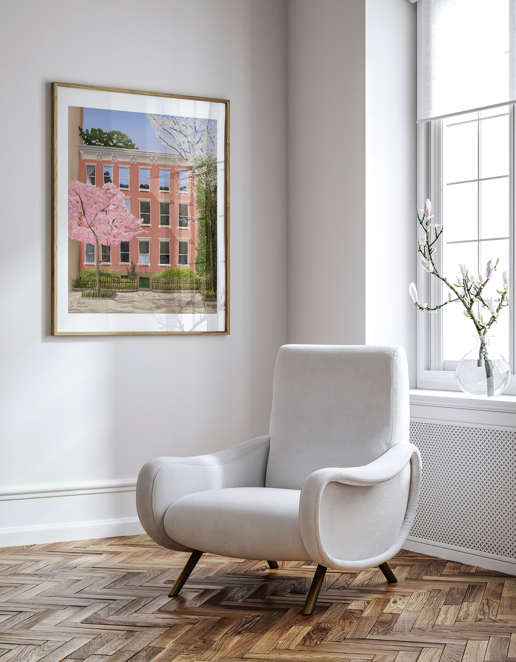 White armchair in a room with a framed picture of a salmon pink building  in the west village and cherry blossoms to the left and a vine on the right, hanging on the wall.