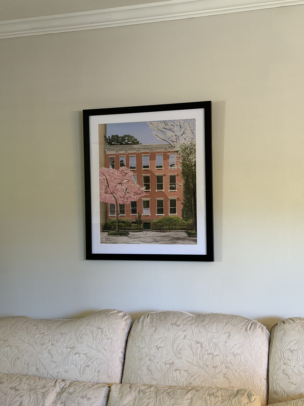 Framed artwork of a brick building with cherry blossoms hanging on a wall above a beige sofa.