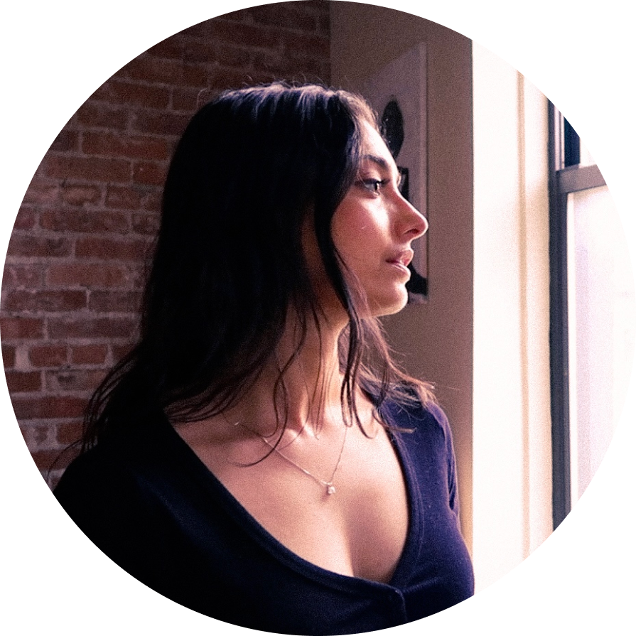 Profile of a long haired brunette woman wearing a v-neck long sleeve black shirt looking toward a window in front of a brick wall with a painting of a woman behind her