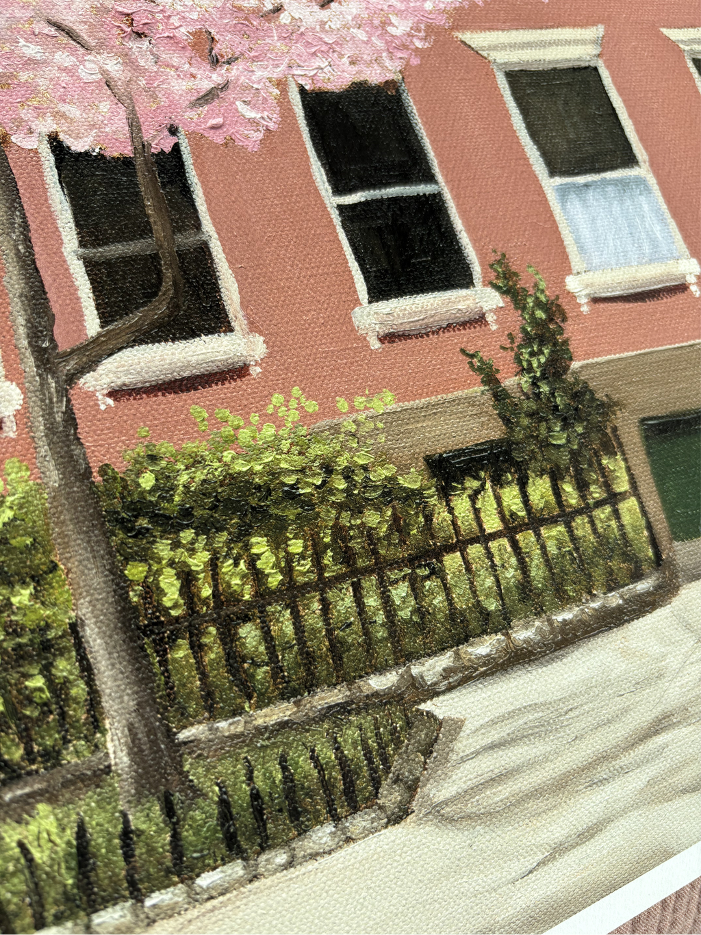 Close up of a painting of a street scene with a cherry blossom tree, red building, and foliage peaking out from the iron fence.