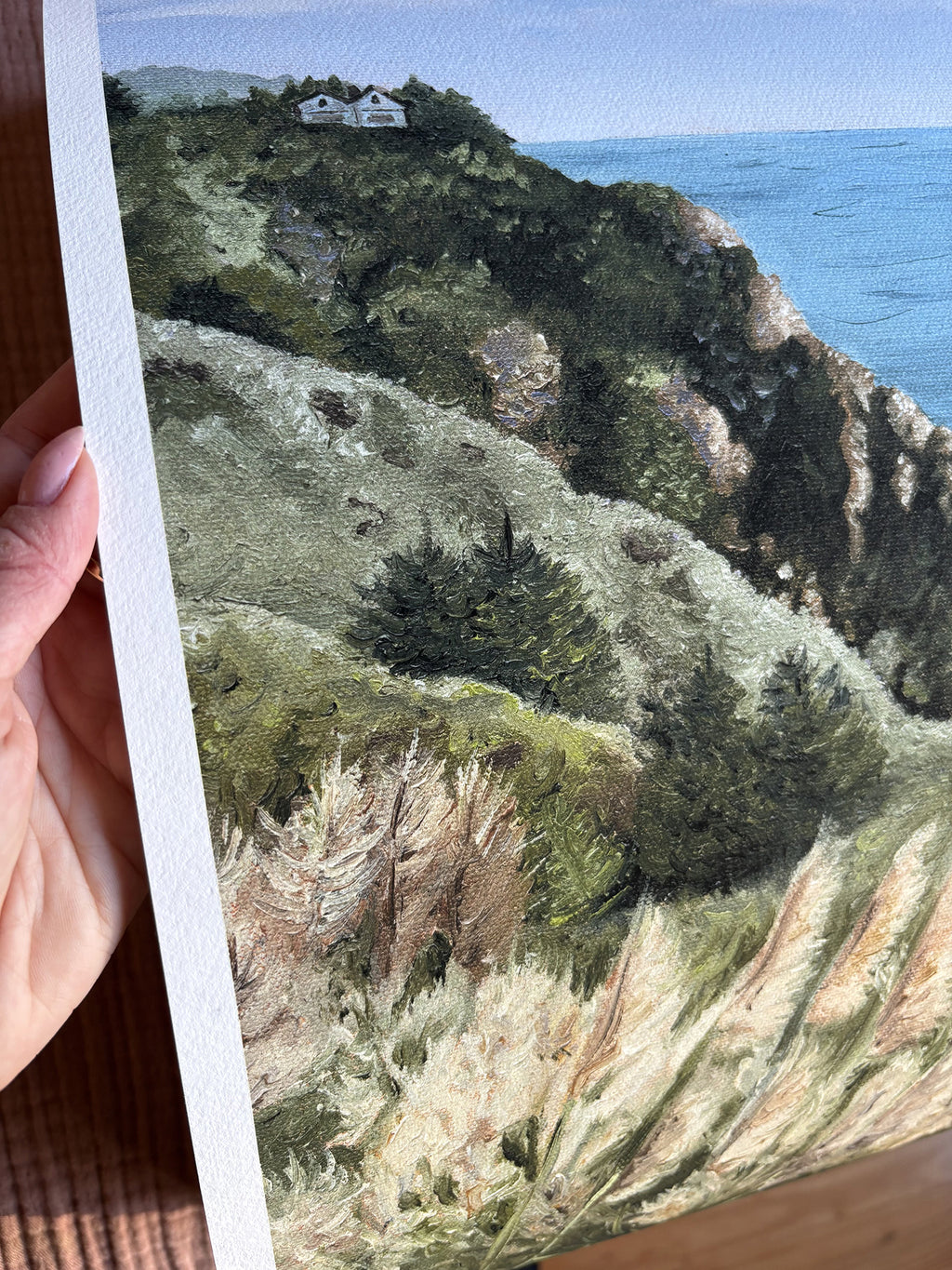 Hand holding a painting of a coastal landscape with a house on a hill.