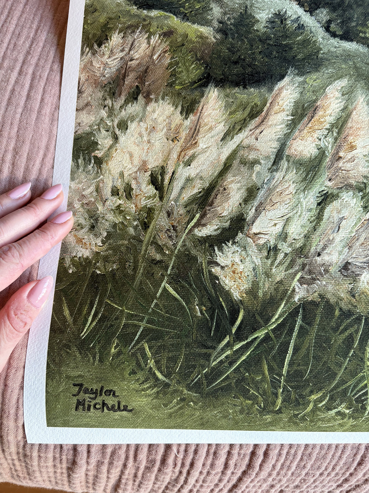 Hand holding a print of a nature scene with reeds and grasses, signed 'Taylor Michele'.