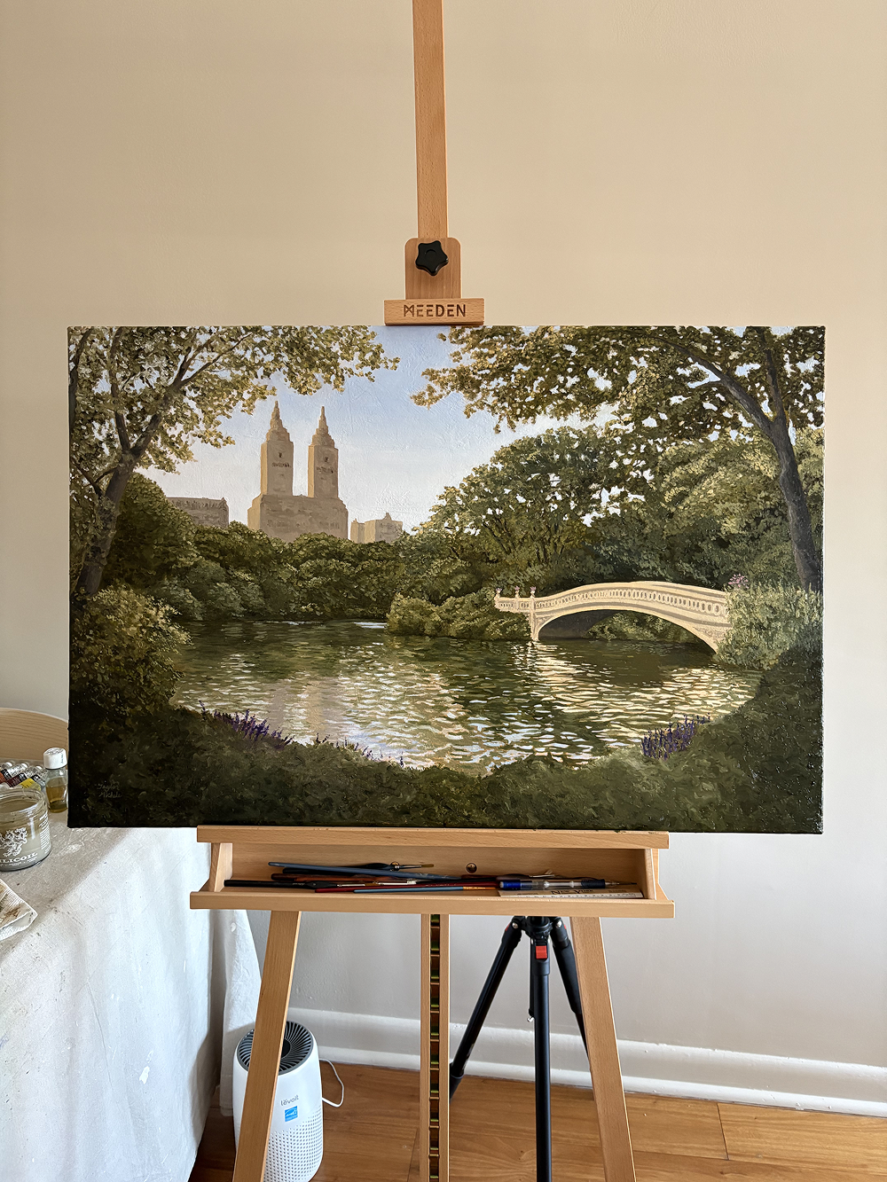 Painting of a park scene with a lake and bridge on an easel.